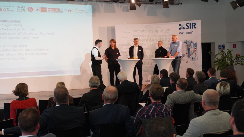 Teilnehmer der Diskussionsrunde auf dem Podium. Zahlreiche Zuhörer sitzen im Publikum.