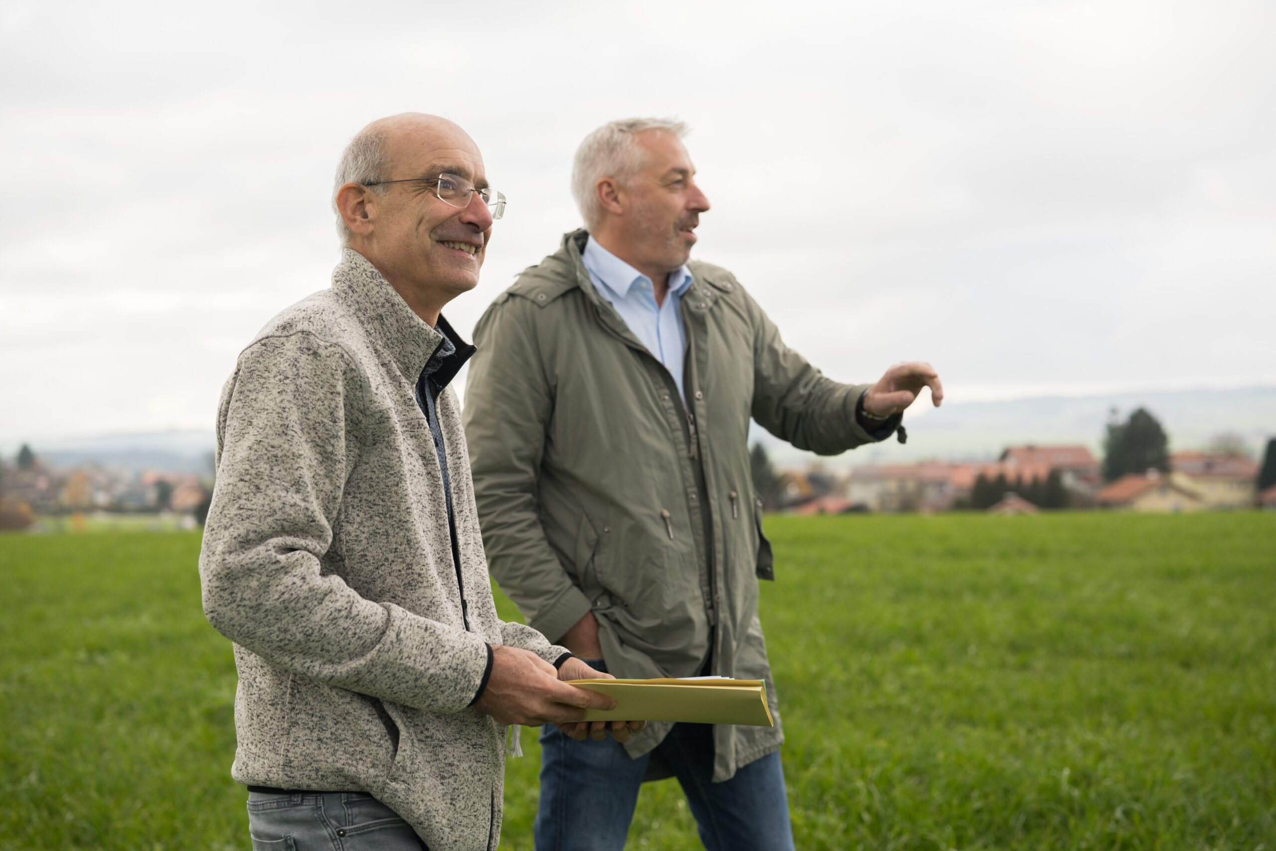 Zwei Mitarbeiter der Land-Invest stehen mit Unterlagen auf einer Wiese. Im Hintergrund ist eine Siedlung zu erkennen.