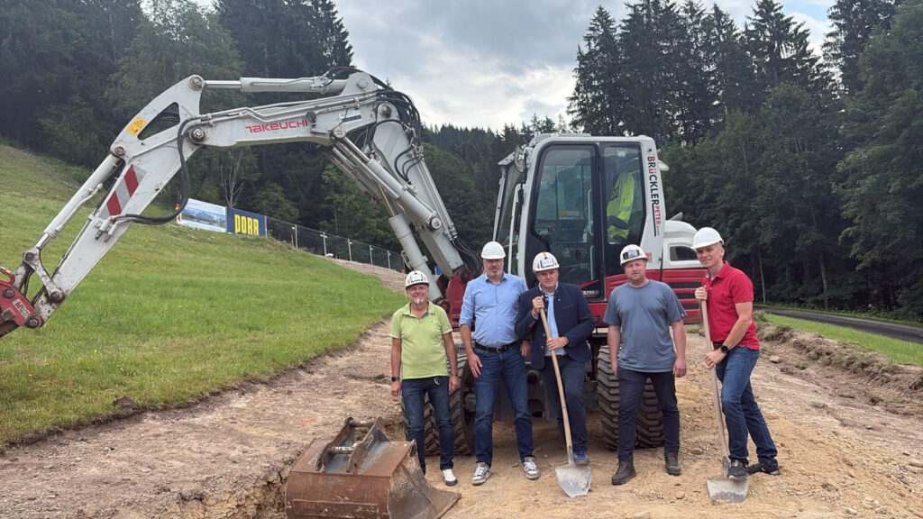 Fünf Männer, in Alltagskleidung aber mit Bauhelmen, stehen vor einem Bagger auf einer Wiese, auf der bereits für eine Straße ein Stück aufgegraben wurde. Die Personen blicken für das Bild in die Kamera - zwei Personen stoßen mit Spaten in den Boden