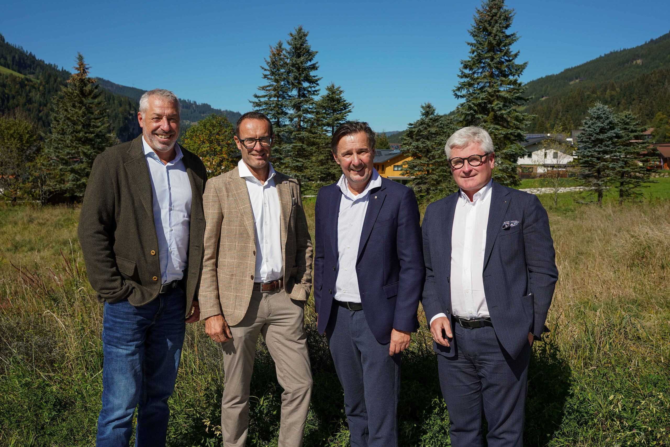 Vier Männer in Sakko stehen freudig lächelnd auf einer hohen Wiese – dem zukünftigen Bauland für das Kleinarler Neubau-Projekt.